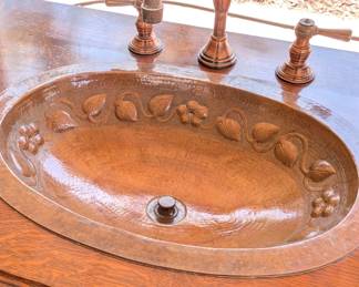 Black Forest style or Dutch Revival hand-carved oak buffet/sideboard that has been custom-converted into a double-sink bathroom vanity. ​The craftsmanship is highly detailed, featuring "Green Man" or mythological face carvings on the frieze and figural scenes on the door panels, likely depicting tavern or village life. The addition of the hammered copper sinks with floral relief and matching oil-rubbed bronze/copper faucets significantly increases its functional value.