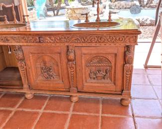 Black Forest style or Dutch Revival hand-carved oak buffet/sideboard that has been custom-converted into a double-sink bathroom vanity. The craftsmanship is highly detailed, featuring "Green Man" or mythological face carvings on the frieze and figural scenes on the door panels, likely depicting tavern or village life. The addition of the hammered copper sinks with floral relief and matching oil-rubbed bronze/copper faucets significantly increases its functional value.