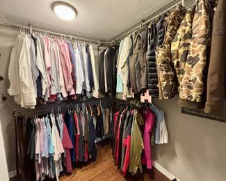 This closet features an extensive collection of men’s clothing organized by style. The upper racks are filled with professional button-down shirts in various colors and patterns, alongside formal blazers and suit jackets. The right side of the rack highlights high-quality outdoor and workwear, including Ariat jackets and several insulated vests in puffer and camouflage styles. The lower racks contain a large assortment of casual polos and short-sleeved shirts, offering a complete range of sizes and styles for both professional and recreational use.