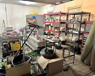 Utility items and professional-grade storage, most notably a classic red Craftsman multi-drawer rolling tool chest and a large diamond-plate aluminum truck bed toolbox. Also pictured are significant home maintenance assets, including a portable pressure washer with a hose reel and multiple large rolls of unused carpeting or turf. The area is organized with various utility shelving units containing various hardware and household supplies.