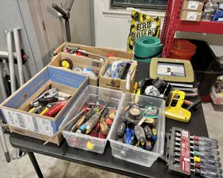 This collection features an extensive variety of hand tools and workshop accessories organized into several bins and boxes for easy browsing. The inventory is headlined by a comprehensive selection of Husky brand tools, including a 7-piece nut driver set and multiple clear bins filled with assorted screwdrivers. Other notable items include several heavy-duty bar clamps, a yellow Stanley staple gun, and various sanding and grinding wheels for power tool attachments. The assortment also contains specialized plumbing shears, hole saws, and decorative picture frames, offering a wide-ranging "handyman’s special" lot of household and workshop hardware