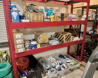 A detailed inventory of contractor-grade building supplies and home improvement hardware. The shelving is stocked with a large volume of LED lighting (Sunperian and Sylvania), including floodlights and interior bulbs, alongside several boxes of new-in-package cabinet and door hardware. The lower level features bulk bins of PVC plumbing fittings and a collection of assorted door knobs, offering a significant opportunity for DIYers or trade professionals looking for stock.