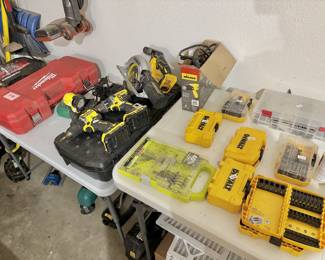 This set includes multiple DeWalt cordless tools, such as a circular saw and two drills with attached battery packs. Notable storage and specialty items include a large red Milwaukee hard carrying case, a Husky mechanics tool set, and a Wagner Furno 300 heat gun in its original box. The table is also lined with various DeWalt and Ryobi drill bit sets and accessory cases, along with a container of wood glue, representing a complete setup for woodworking or construction.