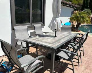 Aluminum 7-piece patio dining set includes a rectangular tile-top table, six sling-back chairs, and a white market umbrella. The Sunvilla Nichols set has four stationary armchairs and two swivel rocker chairs. The table has a wood-look porcelain tile surface and a central umbrella hole. The chairs have a grey weather-resistant sling fabric and dark brown powder-coated aluminum frames.