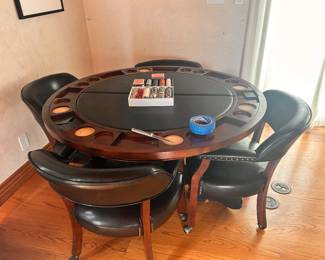 Sturdy wooden poker table set includes four matching black leather upholstered chairs with nailhead trim. The round table has a dark wood finish and a black padded playing surface. The outer rim of the table features built-in cup holders and chip trays for each player. The chairs are equipped with casters for easy movement.