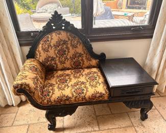 Ornate telephone bench is constructed from dark-stained wood with intricate floral and leaf carvings on the frame, legs, and backrest. The seat and back are upholstered in a tan tapestry fabric featuring a multicolored floral pattern. This piece includes an integrated side table with a single drawer and a curved armrest on the opposite side. We originally bought with $2200, asking for $580