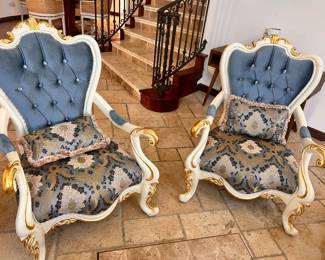 Elegant pair of French-style armchairs with cream-colored wooden frames and gold leaf accents. The backrest is upholstered in tufted blue velvet with crystal-like button detailing. The seat and matching lumbar pillows are covered in a blue and gold damask patterned fabric. Each chair has curved cabriole legs and padded armrests. Need arrange 