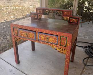 Striking red lacquer Chinese writing desk with ornate gold painted floral and scroll motifs throughout. Features two front drawers with brass hardware and an elevated hutch top with small drawers and carved fretwork. One side unfinished/plain dark wood. Diplomat provenance — a true conversation piece. Approx. 36"W x 20"D x 48"H with hutch.