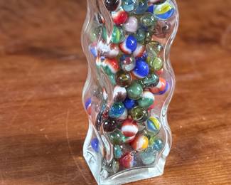 Wavy glass bottle containing a collection of colorful glass marbles. The bottle features a unique wavy design, sealed with a metallic screw cap and says “Roses”. Contents include various multicolored marbles in assorted patterns and designs. Condition appears good with no visible defects to the bottle or marbles.