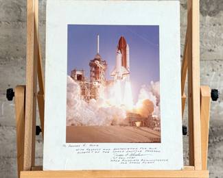 This lot features a collectible vintage NASA photograph depicting a Space Shuttle launch. Signed by NASA associate and measures on backer, 20” H x 16” W.