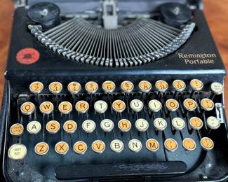 Remington Portable Typewriter featuring a black finish with orange and white round keys. Housed in its original black carrying case with a latch closure. The item shows signs of wear consistent with age and use, including minor scuffs and dust accumulation. Red label detailing model specifications is intact. Includes functioning components but may require overall maintenance or cleaning.