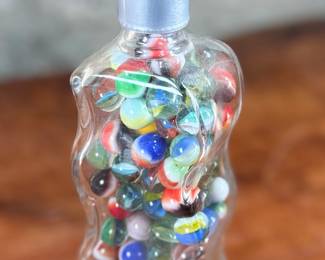 Wavy glass bottle containing a collection of colorful glass marbles. The bottle features a unique wavy design, sealed with a metallic screw cap and says “Roses”. Contents include various multicolored marbles in assorted patterns and designs. Condition appears good with no visible defects to the bottle or marbles.