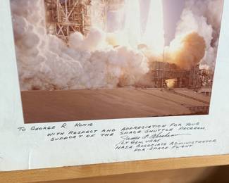 This lot features a collectible vintage NASA photograph depicting a Space Shuttle launch. Signed by NASA associate and measures on backer, 20” H x 16” W.