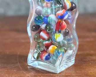 Wavy glass bottle containing a collection of colorful glass marbles. The bottle features a unique wavy design, sealed with a metallic screw cap and says “Roses”. Contents include various multicolored marbles in assorted patterns and designs. Condition appears good with no visible defects to the bottle or marbles.