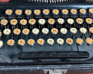 Remington Portable Typewriter featuring a black finish with orange and white round keys. Housed in its original black carrying case with a latch closure. The item shows signs of wear consistent with age and use, including minor scuffs and dust accumulation. Red label detailing model specifications is intact. Includes functioning components but may require overall maintenance or cleaning.