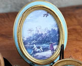 Decorative miniature artworks in various oval and round frames. Frames include painted gold and blue accents, some marked ‘Made in Italy’. Depicting classical European and romantic scenes, including individuals and landscapes. Constructed from materials resembling wood and metal with exposed backing hardware. Intricately set, ideal for wall or tabletop display.