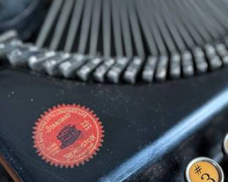 Remington Portable Typewriter featuring a black finish with orange and white round keys. Housed in its original black carrying case with a latch closure. The item shows signs of wear consistent with age and use, including minor scuffs and dust accumulation. Red label detailing model specifications is intact. Includes functioning components but may require overall maintenance or cleaning.