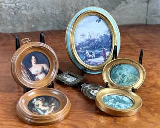 Decorative miniature artworks in various oval and round frames. Frames include painted gold and blue accents, some marked ‘Made in Italy’. Depicting classical European and romantic scenes, including individuals and landscapes. Constructed from materials resembling wood and metal with exposed backing hardware. Intricately set, ideal for wall or tabletop display.
