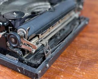 Remington Portable Typewriter featuring a black finish with orange and white round keys. Housed in its original black carrying case with a latch closure. The item shows signs of wear consistent with age and use, including minor scuffs and dust accumulation. Red label detailing model specifications is intact. Includes functioning components but may require overall maintenance or cleaning.