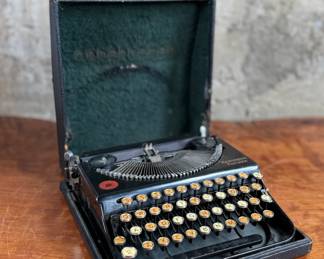 Remington Portable Typewriter featuring a black finish with orange and white round keys. Housed in its original black carrying case with a latch closure. The item shows signs of wear consistent with age and use, including minor scuffs and dust accumulation. Red label detailing model specifications is intact. Includes functioning components but may require overall maintenance or cleaning.