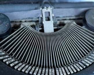 Remington Portable Typewriter featuring a black finish with orange and white round keys. Housed in its original black carrying case with a latch closure. The item shows signs of wear consistent with age and use, including minor scuffs and dust accumulation. Red label detailing model specifications is intact. Includes functioning components but may require overall maintenance or cleaning.