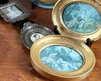 Decorative miniature artworks in various oval and round frames. Frames include painted gold and blue accents, some marked ‘Made in Italy’. Depicting classical European and romantic scenes, including individuals and landscapes. Constructed from materials resembling wood and metal with exposed backing hardware. Intricately set, ideal for wall or tabletop display.