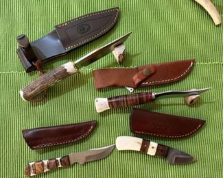 A collection of handcrafted knives featuring stag horn handles and stacked leather handles with multi-colored spacers; one knife is a vintage Randall model with a pinned stag handle and a brown button sheath, another a Marbles Woodcraft knife, which are historic American hunting knives.