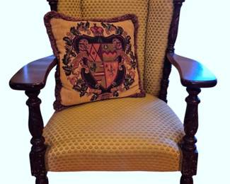609 - Carved Wood Wingback Armchair w/ Pillow (Description): Carved wood wingback armchair featuring  turned legs and arm supports with acorn  finials atop the posts. The chair is  upholstered in a gold textured fabric and  includes a decorative throw pillow with a heraldic coat of arms design. Measures 40" x  29"