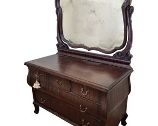 1293 - Carved Mahogany Dresser w/Lady Adorned Mirror (Description): Antique mahogany chest of drawers with  attached mirror. The dresser features a  serpentine bombe-style front with four  drawers fitted with brass bail pulls. The  ornately carved mirror frame displays scrolling acanthus leaf motifs and a  decorative crest. The piece rests on cabriole  legs. Measures 77" high x 51" wide x 25"  deep.