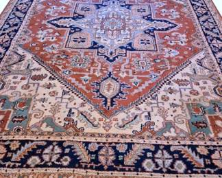605 - Large Heriz Style Oriental Rug 152"x110" (Description): Large Heriz style oriental rug featuring a  classic central medallion design. The rug  displays a striking geometric medallion in  shades of blue and navy against a rich  rust-red field. Cream corner spandrels contain traditional floral and botanical  motifs in teal, orange, and earth tones. A  wide navy border surrounds the field with  repeating palmette and leaf patterns. The rug  features hand-knotted wool construction with