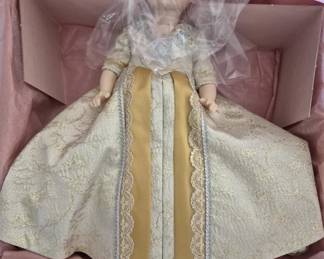 1384 - Madame Alexander Mary McKee doll 13" (Description): Madame Alexander Mary McKee doll measuring 13  inches. The doll is presented in its  original box with pink tissue paper,  featuring an elegant gold and cream brocade  gown with lace trim accents. The doll has blonde hair and comes with protective plastic  covering over the head.