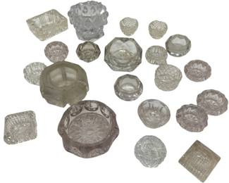989 - Collection of Pressed Glass Salt Cellars (Description): A collection of pressed glass salt cellars in  various shapes and sizes. The assortment  includes square, round, and decorative forms  featuring different cut glass patterns  including diamond, starburst, and geometric designs. Some pieces show a subtle amethyst  tint indicating age, while others are clear  crystal glass. The collection represents  various styles typical of Victorian and early  20th century tableware.