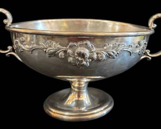 531 - Sheffield Plated Trophy Bowl 6"x14.5" (Description): Sheffield plated trophy bowl featuring  elegant double scroll handles and a footed  pedestal base. The bowl is decorated with a  repousse floral and foliate band around the  exterior, showcasing a central flower motif flanked by ornate acanthus leaves. The piece  displays classic Victorian styling with its  graceful curved handles and refined  proportions. Measures 6 inches by 14.5  inches.