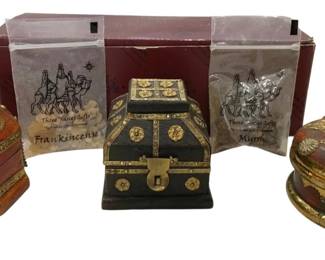 977 - Three Kings Gifts Frankincense & Myrrh Set (Description): A decorative Three Kings Gifts set featuring  three ornate treasure chest-style boxes  containing frankincense and myrrh. The set  includes three distinctly styled boxes: one  hexagonal red and gold box, one black and gold rectangular chest, and one round red and  gold lidded container. Each box features  detailed metallic embellishments and antiqued  finishes. The set comes in original  packaging depicting the three wise men on