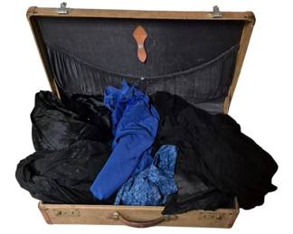 4101 - Vtg Suitcase w/ Assorted Vtg Clothing (Description): A vintage suitcase containing a collection of  vintage clothing. The suitcase features a  tan tweed exterior with brown leather trim  and brass hardware. Inside, the lot includes  various garments in black and blue tones, including what appears to be blue fabric  pieces and dark fabric items. The suitcase  interior is lined in black fabric with a  leather strap detail on the lid.