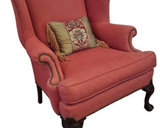 310 - Coral Upholstered Wing Back Chair 42"x34"x28" (Description): A classic wing back chair upholstered in  coral pink fabric with brass nailhead trim  along the rolled arms. The chair features a  gracefully curved camel back and rests on  carved dark wood cabriole legs with ball and claw feet in the Queen Anne style.
