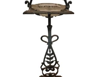 3750 - Iron Ash Tray / Smoking Stand 26.5" (Description): Decorative iron smoking stand with ash tray,  measuring 26.5 inches tall. The stand  features an ornate wrought iron design with  scrollwork and a leaf motif on the central  support. The base is a substantial square pedestal with architectural details. The top  includes a glass ash tray insert set within  an ornamental iron frame with a curved  carrying handle. The piece exhibits a dark  patina consistent with vintage ironwork.