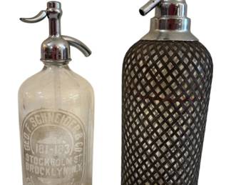 572 - 2 Vtg Seltzer Bottles Geo. F. Schneider & Co (Description): Two vintage seltzer bottles measuring 13.5  inches tall. One is a clear glass bottle with  etched advertising for Geo. F. Schneider &  Co., 181-183 Stockholm St., Brooklyn, N.Y.,  featuring a chrome siphon top. The second is a glass seltzer bottle encased in a wire mesh  protective covering with a chrome  double-spout dispenser top.