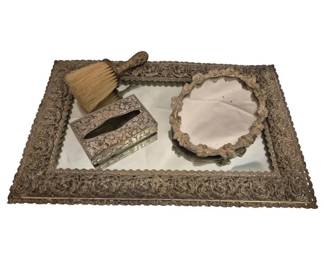 2740 - Vtg Ornate Brass Vanity Set w/ Mirrored Tray (Description): Vintage vanity accessories featuring an  ornate brass mirrored tray with elaborate  scrollwork border and scalloped edge. The set  includes a decorative tissue box cover with  matching repousse design, a natural bristle clothes brush with ornate handle,and a small  round hand mirror with floral frame. The  pieces display Victorian-inspired styling  with intricate embossed patterns throughout.