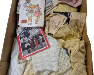 4132 - Assorted Doll Clothes & Sewing Patterns (Description): A collection of assorted doll clothes and  vintage sewing patterns. The lot includes  handmade crocheted and knitted items in  white, cream, and ivory tones, along with  various vintage sewing patterns including Simplicity patterns for toddlers' smocked  dresses and children's clothing. Also  included are lace trims, fabric pieces in  pink, and additional crafting materials.