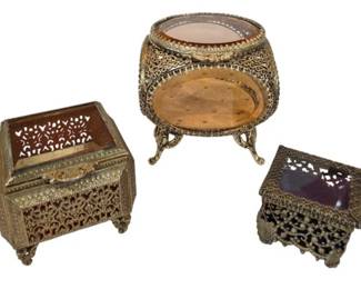 2714 - 3 Vintage Gilt Filigree Jewelry Boxes (Description): Three vintage jewelry boxes featuring ornate  gilt filigree metalwork. The collection  includes a rectangular chest-style box with  hinged lid, an oval footed casket with  beveled glass top, and a smaller square trinket box. Each piece displays intricate  pierced decorative patterns in the  ormolu-style metalwork with glass viewing  panels, characteristic of mid-century vanity  accessories.