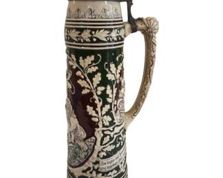 551 - German Stoneware Beer Stein with Pewter Lid (Description): Large German stoneware beer stein featuring a  pewter lid with pointed finial. The stein is  decorated with relief oak leaf and acorn  motifs in green, burgundy, and cream colors.  A hunting scene with deer is depicted on the body, and German text adorns the base. The  ornate handle features detailed scrollwork.  Measures 19 inches tall by 5.5 inches wide.