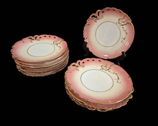 488 - 12 Limoges Pink & Gold Plates 8.5" (Description): Set of twelve Limoges porcelain plates  featuring a distinctive pink ombre border  graduating to a cream center. Each plate is  embellished with elegant gold scrollwork  accents and gilded scalloped edges. The plates display a Rococo-inspired design with  pierced decorative handles at the top.  Measuring 8.5 inches in diameter, this set  exemplifies the refined French porcelain  craftsmanship for which Limoges is renowned.