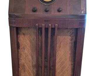 3723 - Philco Model 38-4 Vintage Floor Radio (Description): Philco Vintage Radio model 38-4 in a wooden  floor console cabinet. This Art Deco era  radio features a distinctive round dial face  at the top with tuning controls beneath, and  a large fabric-covered speaker grille with vertical wooden bars on the front. The  cabinet displays attractive wood grain veneer  panels. Measures 42 inches wide by 25 inches  tall by 16 inches deep. Used as is electric/electronic/mechanical
