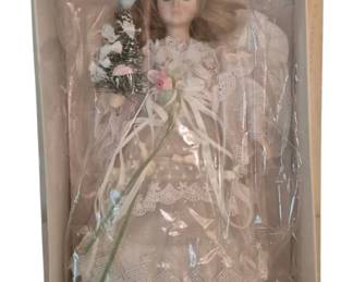 4062 - Kurt Adler Angel Tree Topper Original Box (Description): Vintage Kurt Adler angel tree topper in its  original box. This elegant angel features a  porcelain head with delicate blue eyes and  flowing auburn hair adorned with a floral  wreath. She wears an elaborate white lace gown with intricate embroidery and scalloped  trim, and holds a bouquet of silk flowers  with trailing ribbons. The angel has ethereal  white wings and retains the original plastic  covering within the display box.
