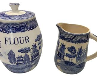 982 - Blue Willow Flour Canister & Pitcher Set (Description): Both pieces feature the classic transferware  design with traditional Chinese-inspired  motifs including pagodas, trees, and  landscape scenes in cobalt blue on a white  ground. The barrel-shaped canister is marked "FLOUR" in blue lettering and include a  fitted lid with knob finial. The pitcher  features a comfortable handle and  coordinating Blue Willow decoration. Measures  9.5" x 6".