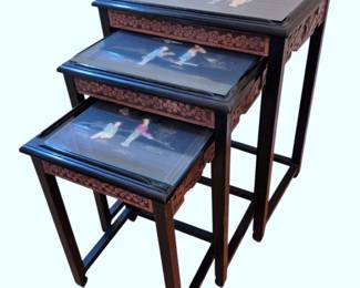 601 - Chinese Black Lacquer Nesting Tables Set of 3 (Description): Set of three Chinese black lacquer nesting  tables featuring hand-painted figural scenes  on the glass-topped surfaces depicting  traditional figures in a landscape setting.  The tables showcase carved floral motifs along the apron edges in a contrasting  reddish-brown tone against the black lacquer  finish. Each table is supported by straight  legs with stretcher bases in classic Chinese  style. Measurements: 26"x20"x14".
