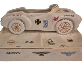 3783 - Ceramic Automobile Dresser Tray 3"x7.5"x5" (Description): Ceramic automobile dresser tray featuring a  vintage race car atop a rectangular base. The  piece is decorated with numerous classic  automotive manufacturer logos and emblems  including Ferrari, MG, Rolls Royce, Singer, Cooper, Austin, and Bentley among others. The  cream-colored ceramic has gilt trim accents.