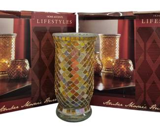4025 - 2 Sets of 3 Amber Mosaic Hurricane Candles (Description): Two boxed sets of Home Accents Lifestyles  Amber Mosaic Hurricanes, each set containing  three pieces. The mosaic glass hurricanes  feature an iridescent amber and gold diamond  pattern design. One hurricane is displayed outside the boxes, showcasing the decorative  mosaic tile work in warm golden tones. Sets  remain in original retail packaging.