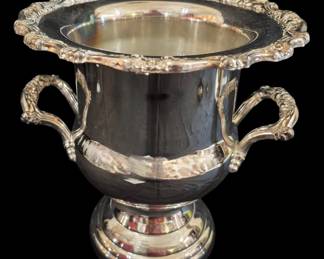 518 - Oneida Silver Plated Champagne Bucket 10" (Description): Oneida silver plated champagne bucket  featuring an elegant urn-form design with a  flared rim adorned with ornate grape and vine  motifs. The bucket is fitted with twin  scrolled handles decorated with matching grapevine detailing, resting on a stepped  circular pedestal base. This classic piece  exemplifies traditional barware styling.  Measures 10 inches tall.